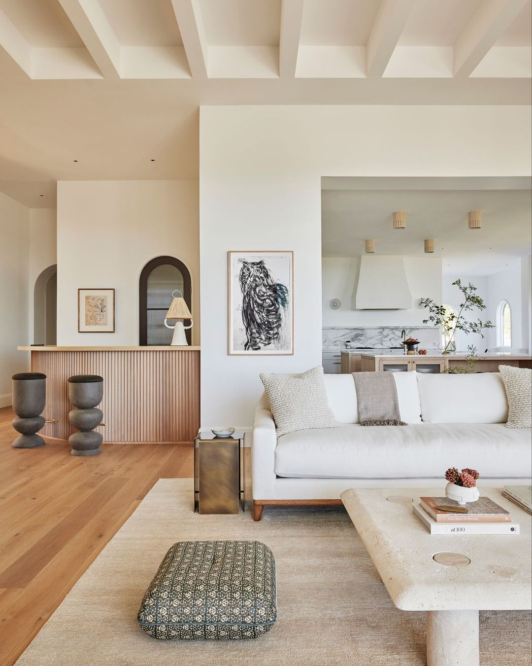 Bright, modern living room features a white sofa, patterned ottoman, light wood floors, and neutral decor with contemporary artwork.