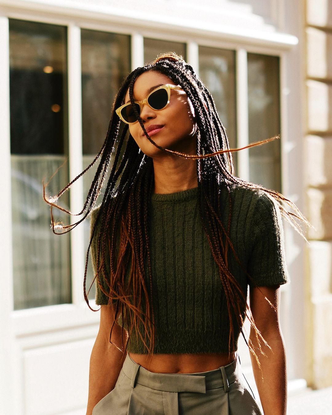 A young woman with long braids poses in an olive-green crop top and high-waisted trousers against a sunlit, neutral building facade.