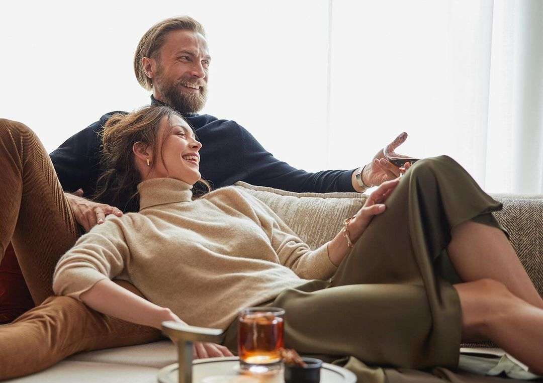 A casually dressed couple lounges together on a neutral sofa in a bright indoor space, drinking whiskey and smiling.