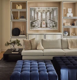 Modern living room interior featuring a large light grey sofa, two blue velvet ottomans, and built-in shelving with decorative objects.