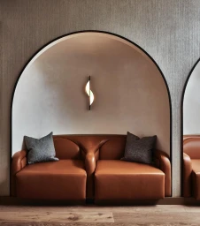 Tan leather sofa inside an arched alcove with two grey pillows and a modern vertical wall sconce