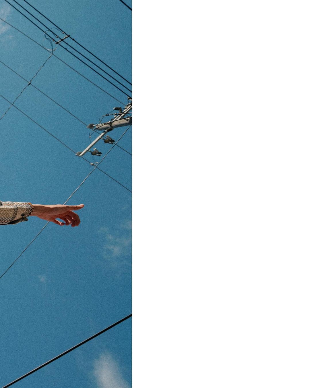An arm extends pointing upwards to a bright blue sky with power lines, showcasing a minimalist composition under natural sunlight.