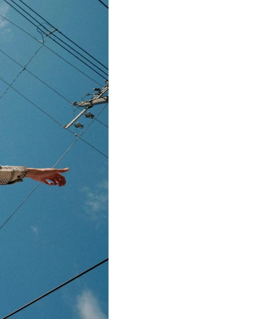An arm extends pointing upwards to a bright blue sky with power lines, showcasing a minimalist composition under natural sunlight.