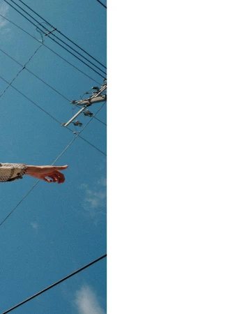 An arm extends pointing upwards to a bright blue sky with power lines, showcasing a minimalist composition under natural sunlight.
