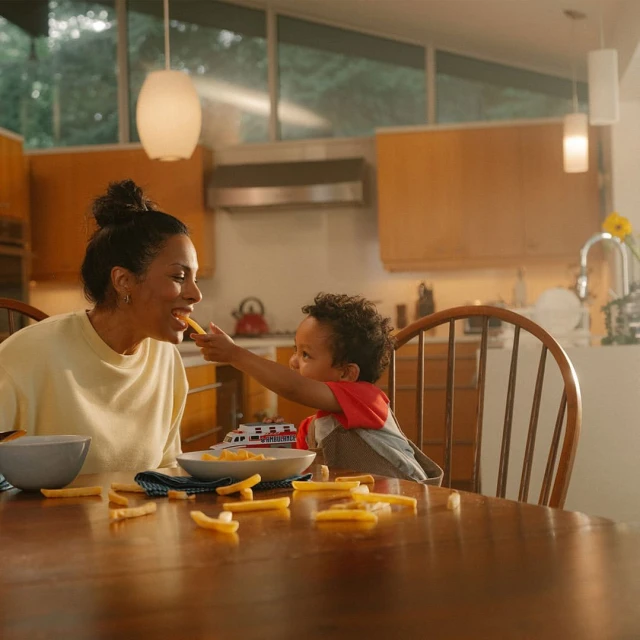 A young child feeds french fries to an adult female sitting at a wooden table inside a home with a view of trees outside.