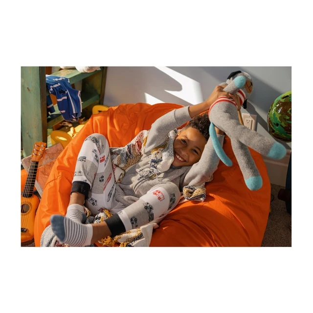Smiling young boy lounges on an orange beanbag chair, playfully holding a grey sock monkey toy in an indoor setting with a ukulele.