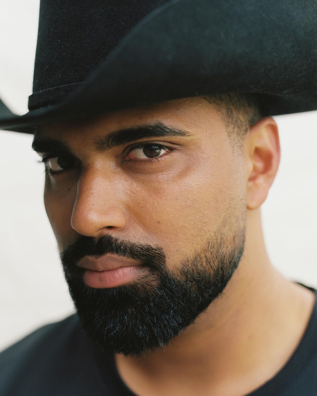 Close-up portrait of a Middle Eastern adult male with a beard wearing a black cowboy hat against a plain white backdrop.