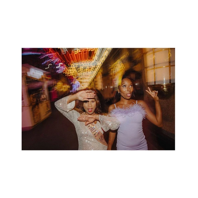 Two young women in dresses pose with hand gestures, illuminated by bright neon lights at night in an urban setting with motion blur.