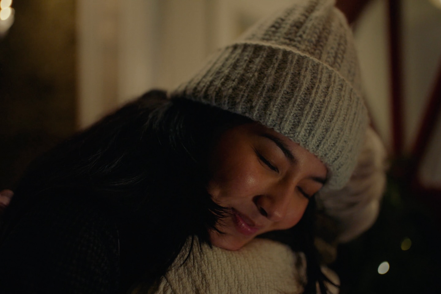 Close-up of a smiling young woman wearing a grey beanie hugging someone tightly, with Christmas tree lights softly blurred in the background.