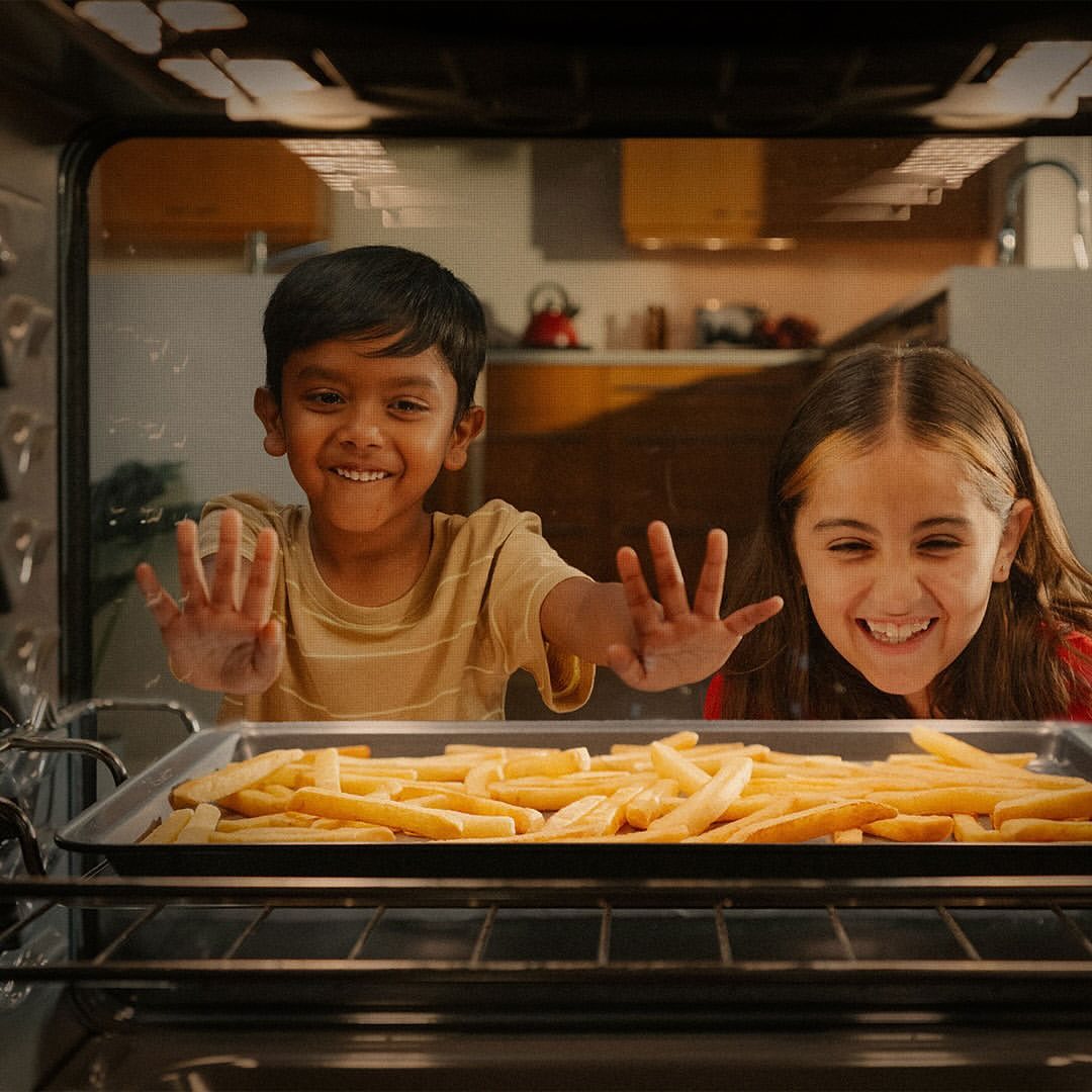 Two children are excitedly looking into an oven at a tray of french fries, indoor kitchen setting, medium shot, warm lighting.