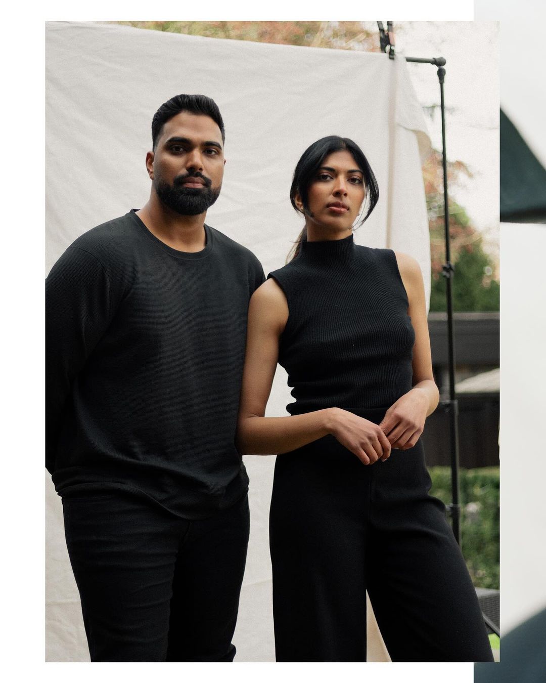 Portrait of a South Asian man and woman in black apparel standing together against a white fabric backdrop.