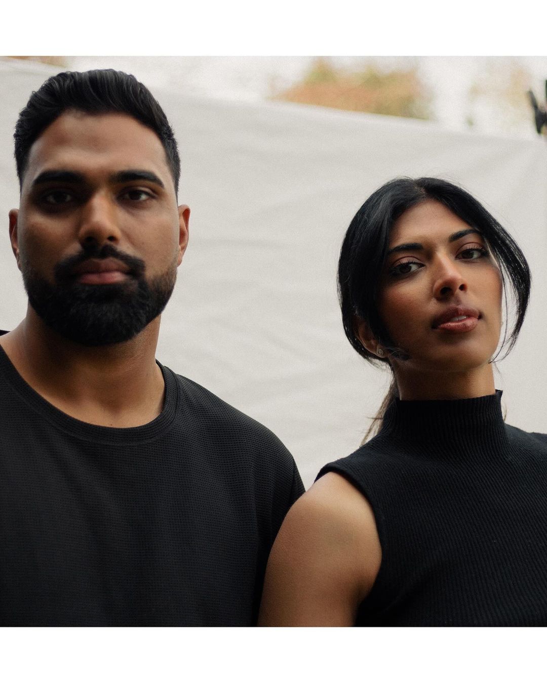 Portrait of a South Asian couple against a white backdrop. The male has a beard and black sweater, and the female has a sleeveless top.