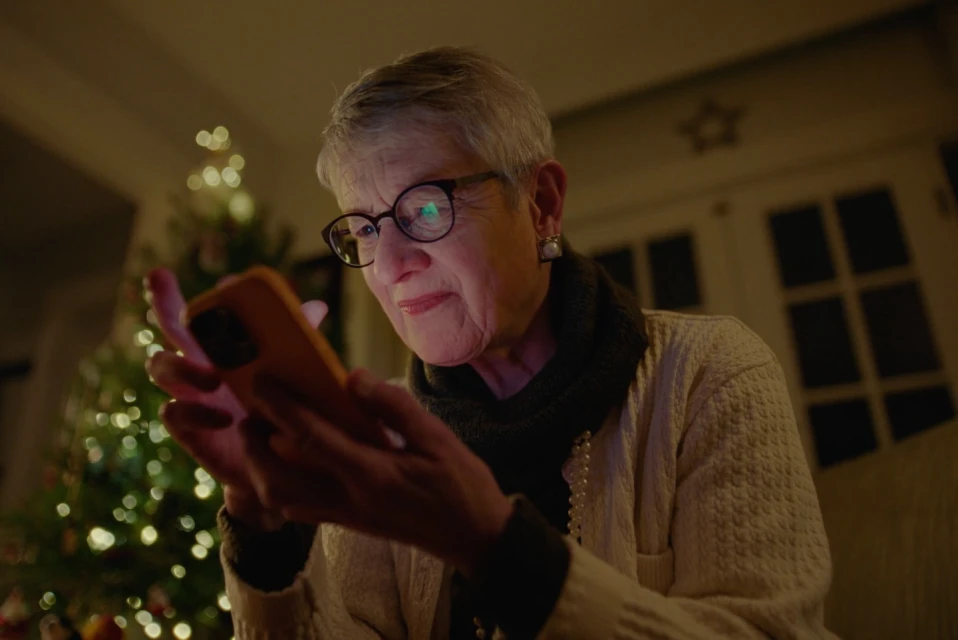 Elderly woman with short gray hair, wearing glasses, is illuminated by a phone screen in a softly lit room with Christmas tree.