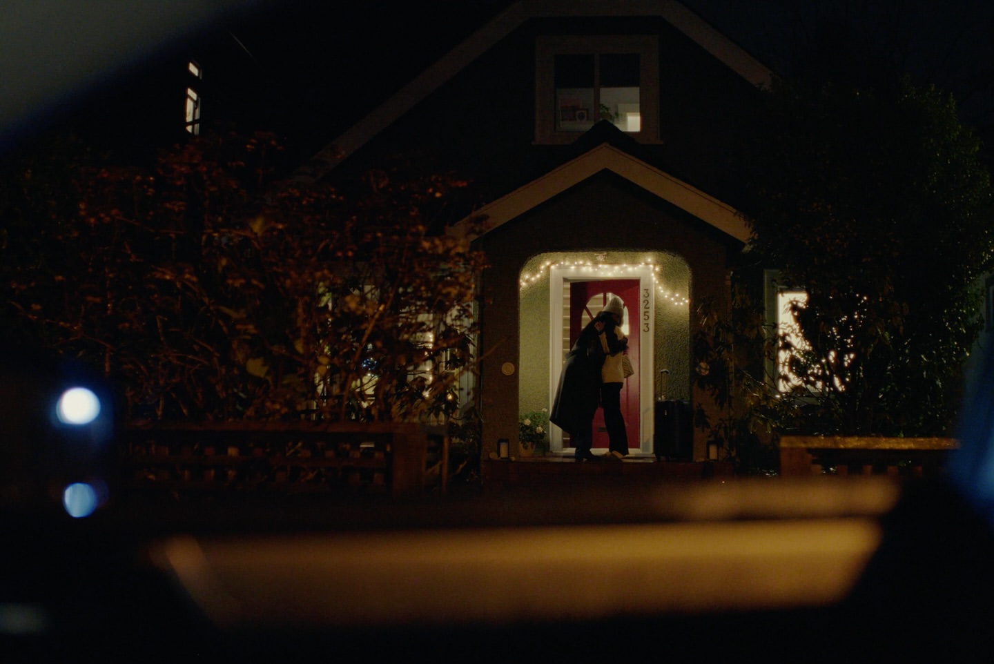 Two people embracing outside a house with a red door and string lights at night, viewed from inside a car through the front windshield.