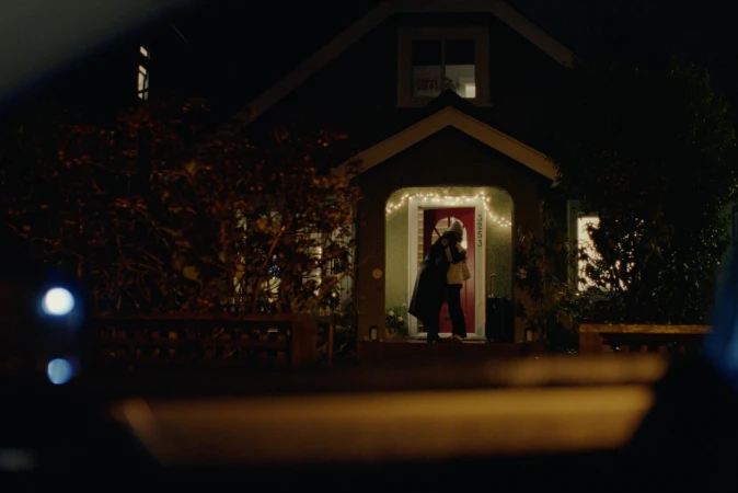 Two people embracing outside a house with a red door and string lights at night, viewed from inside a car through the front windshield.