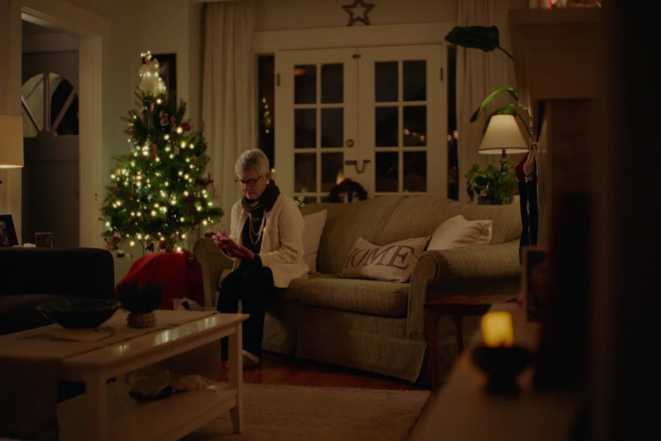 Elderly woman sits on a couch in a cozy living room near a Christmas tree, holding a gift, creating a warm, holiday atmosphere.