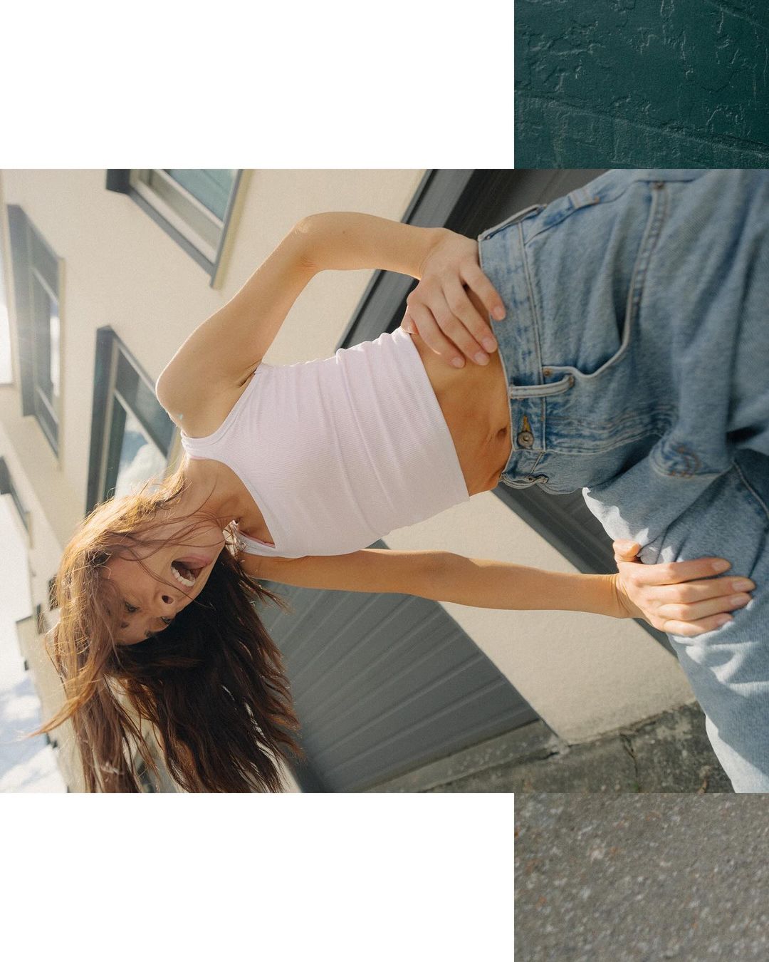Low-angle shot of a young adult female with long brown hair wearing a white tank top and jeans, posing outdoors with a building behind her.