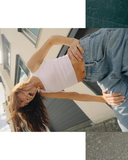 Low-angle shot of a young adult female with long brown hair wearing a white tank top and jeans, posing outdoors with a building behind her.