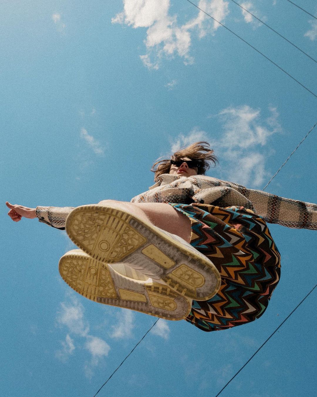 Low-angle action shot of a person jumping against a bright blue sky, showing patterned sneakers and zigzag skirt.