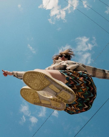 Low-angle action shot of a person jumping against a bright blue sky, showing patterned sneakers and zigzag skirt.