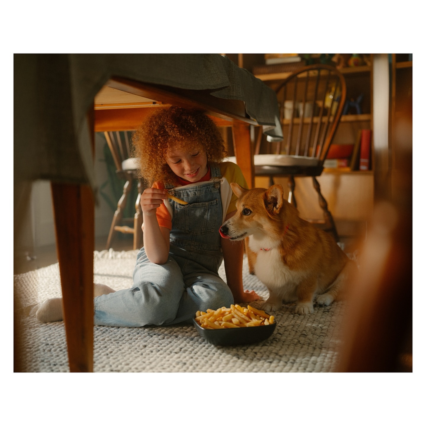 Child and corgi dog share french fries under a table. The girl has curly red hair and wears overalls. Indoor scene with natural light.