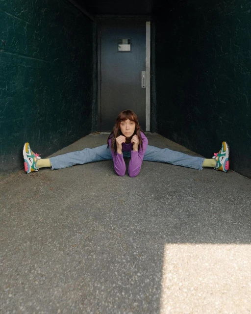 Adult female in a split pose, wearing a purple long-sleeved shirt, blue jeans, and multicolored sneakers in a green-walled corridor.