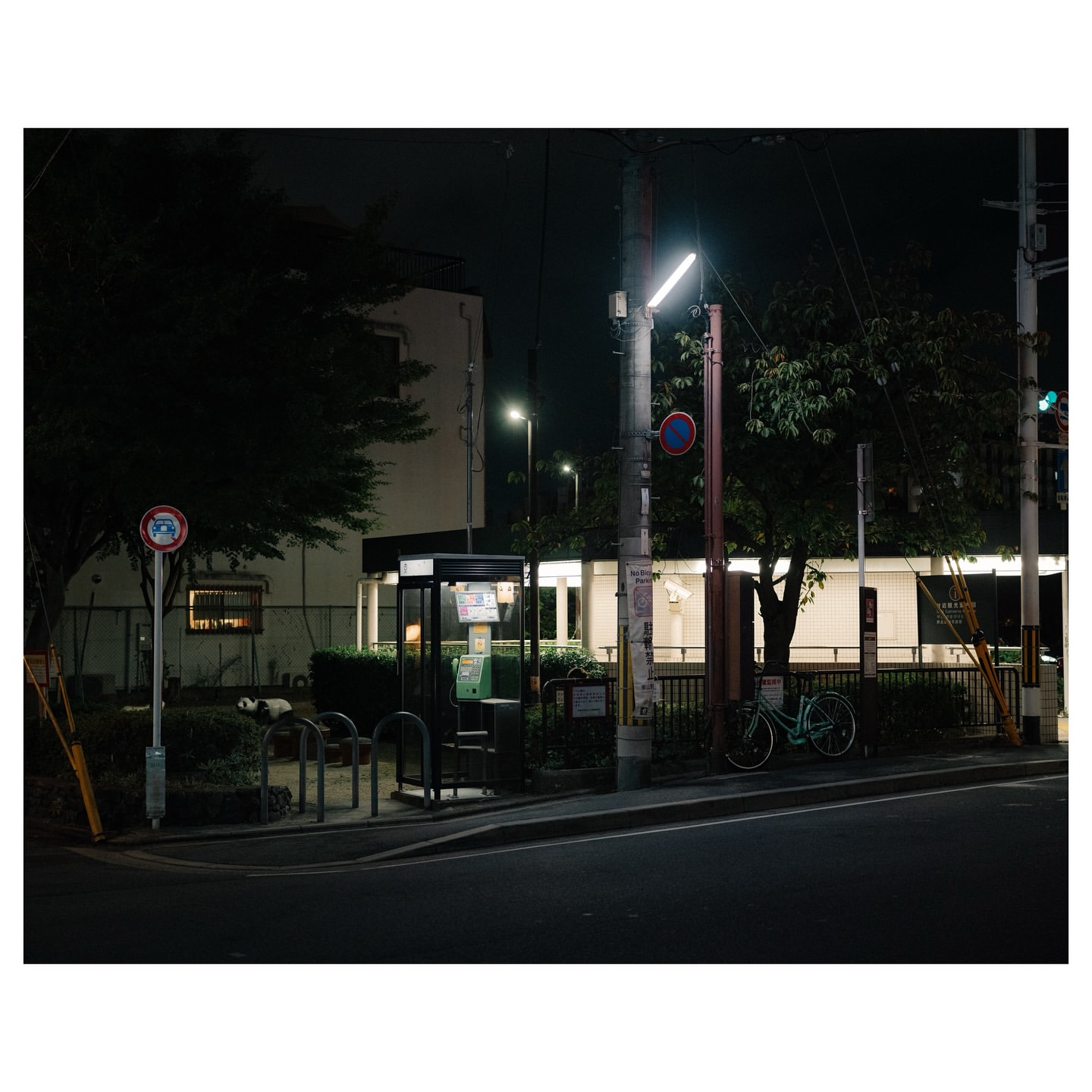 Night scene featuring a bicycle parked near a bus stop and telephone booth, with surrounding foliage and building in the background.