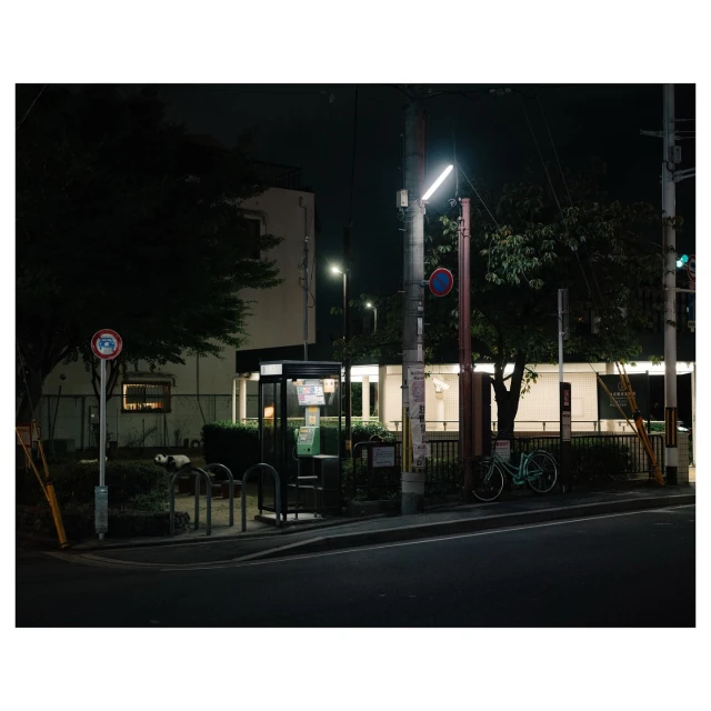 Night scene featuring a bicycle parked near a bus stop and telephone booth, with surrounding foliage and building in the background.
