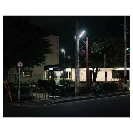 Night scene featuring a bicycle parked near a bus stop and telephone booth, with surrounding foliage and building in the background.