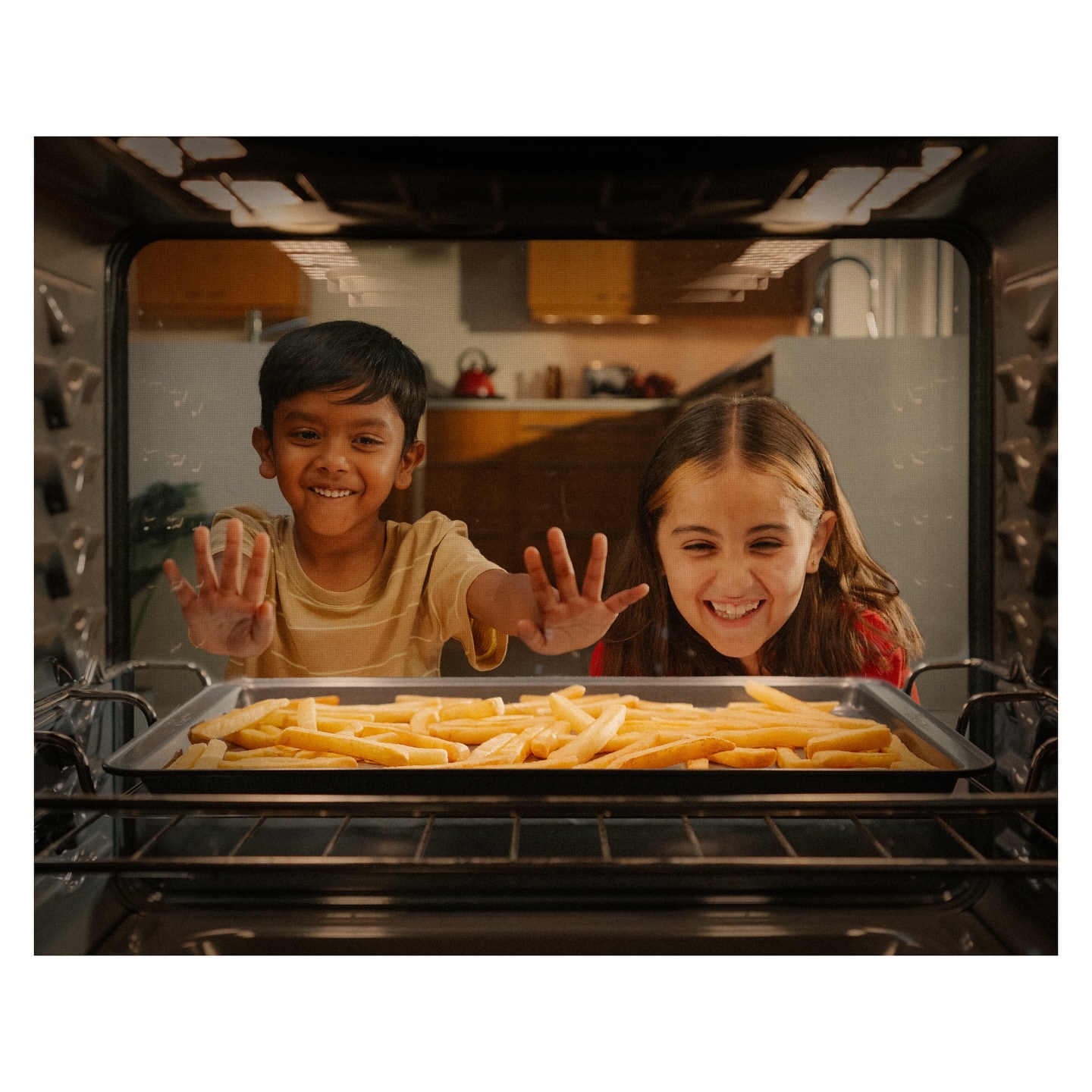 Two smiling children look into an oven filled with french fries on a baking sheet in a brightly lit kitchen, both with hands raised.