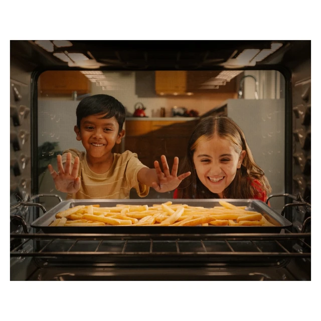 Two smiling children look into an oven filled with french fries on a baking sheet in a brightly lit kitchen, both with hands raised.