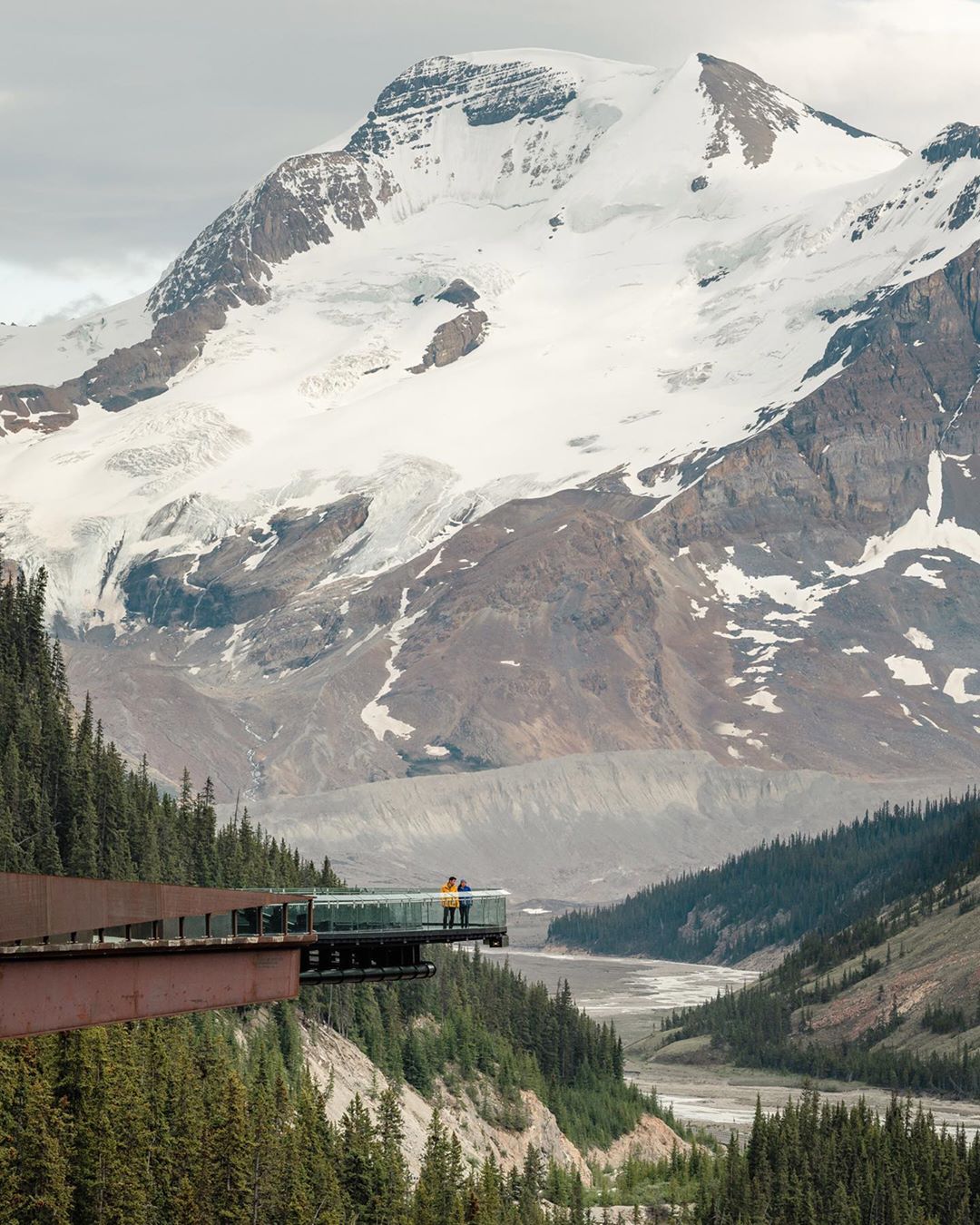 Two people stand on the glass-floored Glacier Skywalk in Jasper National Park, Alberta, with snow-covered mountains in the background.