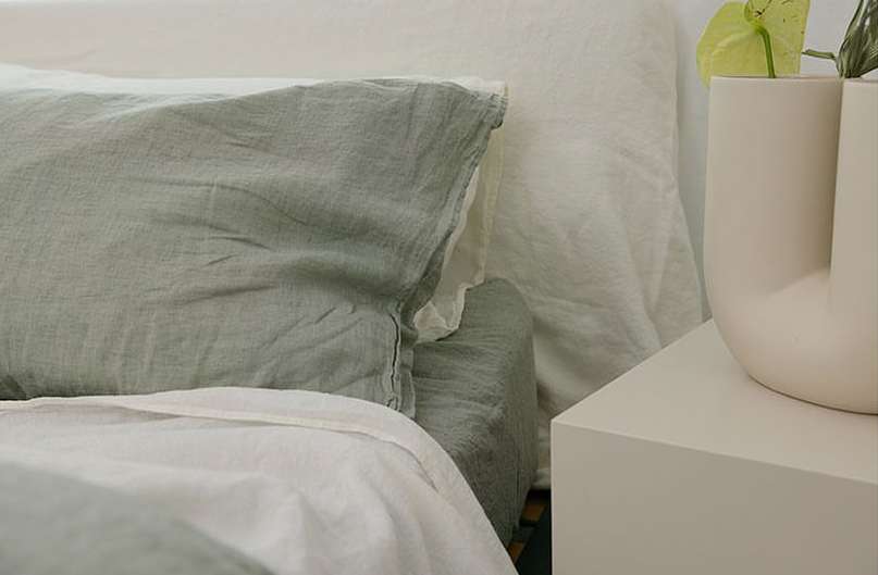 Tranquil close-up showcases a sage green pillow and white bedding in a serene bedroom, complemented by a minimalist white vase and plant.