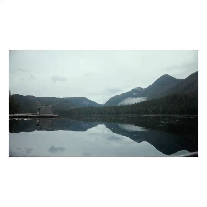 Scenic landscape featuring a tranquil lake reflecting forested mountains under a cloudy sky, with a small boathouse on the water's edge.