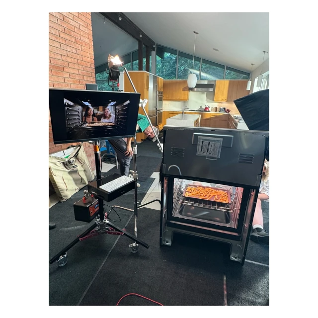 Behind-the-scenes look at a filming set featuring a monitor displaying two women, kitchen appliances, studio lighting, and camera equipment.