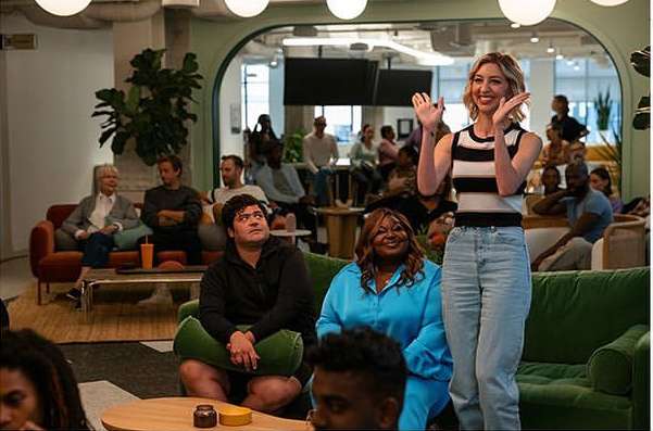 Indoor shot of a smiling woman in a striped top and jeans standing with raised hands in front of a seated audience in a lounge area.