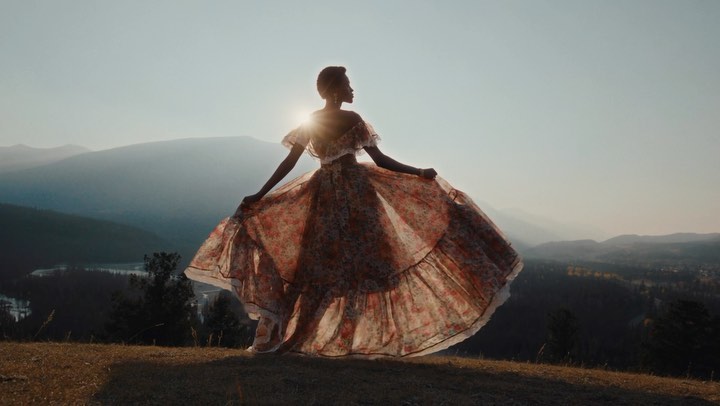 An adult female in a floral dress poses on a hilltop against a mountain background with sun flare. The woman's dress flows in the breeze.
