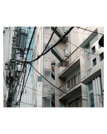 Urban scene featuring a man on a balcony of a building with exposed fire escapes and a utility pole with tangled wires.