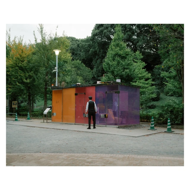 Man in waistcoat and hat stands facing colorful transparent public restrooms in park setting with green trees and cloudy sky.
