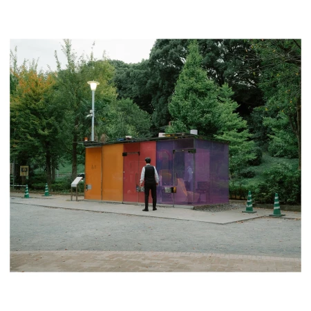 Man in waistcoat and hat stands facing colorful transparent public restrooms in park setting with green trees and cloudy sky.