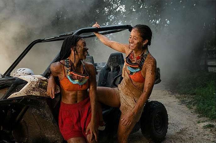 Two women covered in mud pose with an off-road vehicle in a rural setting, surrounded by trees and smoke, after an adventurous ride.