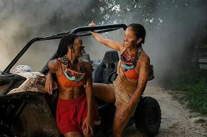 Two women covered in mud pose with an off-road vehicle in a rural setting, surrounded by trees and smoke, after an adventurous ride.