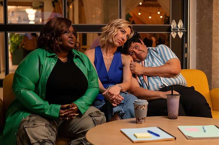 Three adults sitting closely together on a yellow sofa inside a modern office setting with visible natural light from nearby windows.