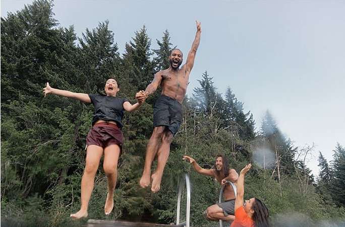 A group of young adults jumps into a pool, captured from a low angle with a background of lush green trees and a clear blue sky.