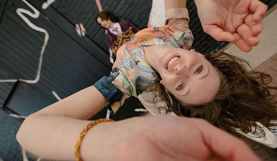 Upside-down eye-level shot of smiling woman in patterned top with arm raised, indoor setting, another person lying in background.