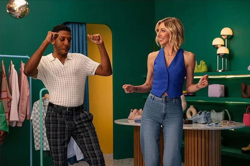 Two adult people dancing in a brightly colored retail or studio space, with clothing racks, shelves of accessories, and creative lamps.
