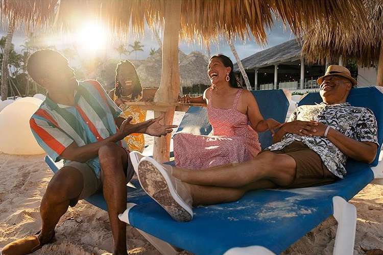 A group of four people sitting and laughing on a beach under a thatched umbrella near a resort on a sunny day with the ocean visible.