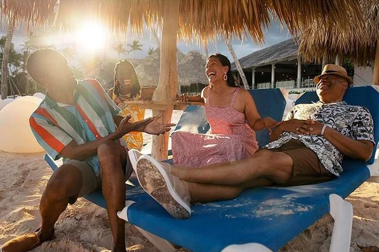 A group of four people sitting and laughing on a beach under a thatched umbrella near a resort on a sunny day with the ocean visible.