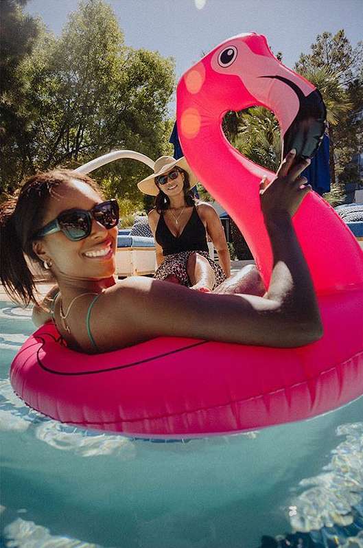 Two women relax in a pool; one in a flamingo floatie, the other on a lounge chair, enjoying the sunshine on a summer day.