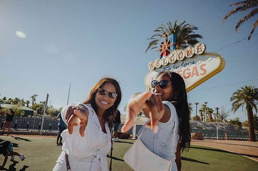 Two smiling women pose in front of the 'Welcome to Fabulous Las Vegas' sign, both wearing sunglasses and casual summer outfits on a sunny day.